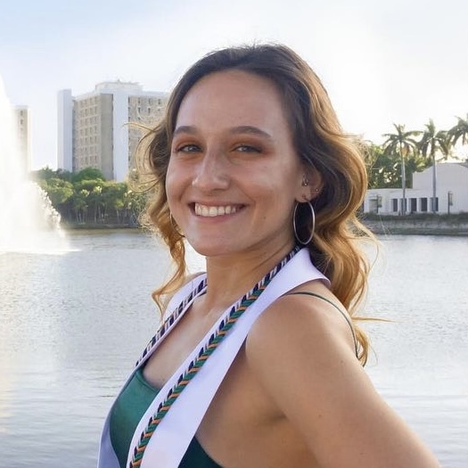 Picture of a smiling person with long brown curled hair, fair skin, and a white sash around their neck and a green dress with a blue sky, lake and buildings in the background.