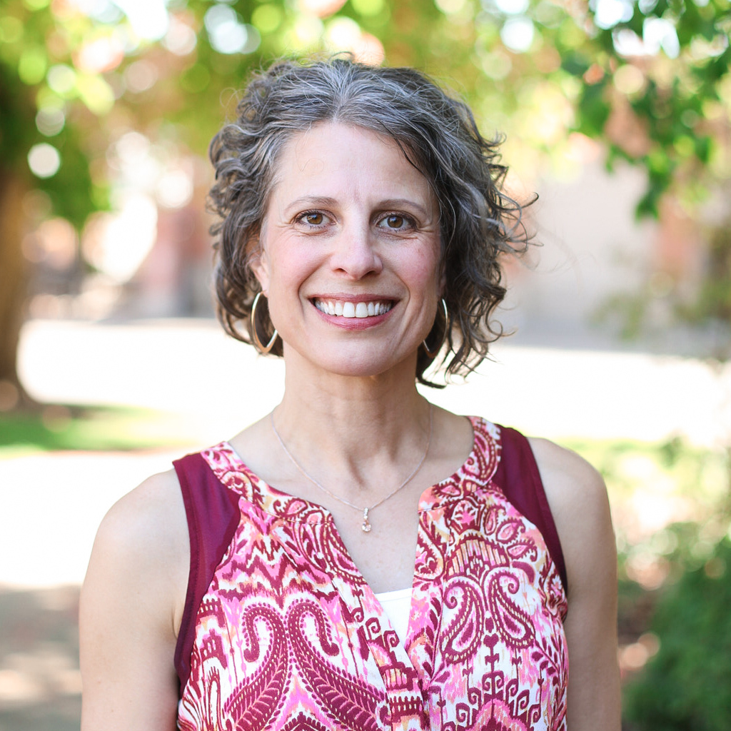 Selfie of a person with medium length curly hair, fair skin, and a black rain jacket against a background full of trees.