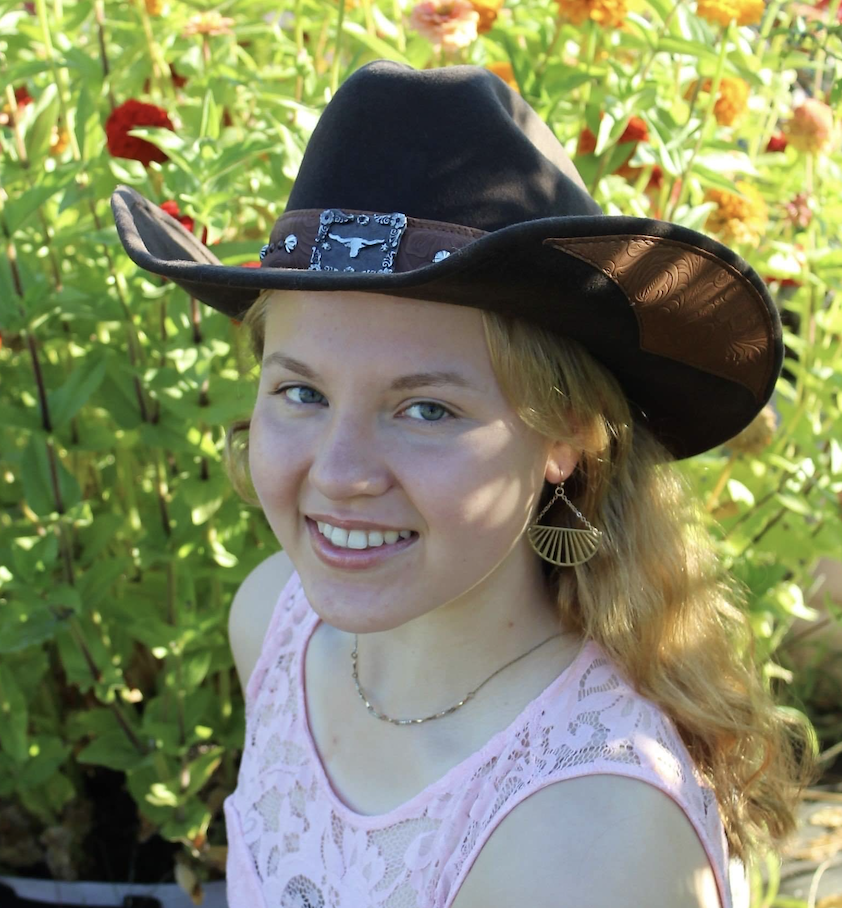 Photo of a person with blond curly hair wearing a brown cowboy hat against a backdrop of greenery.