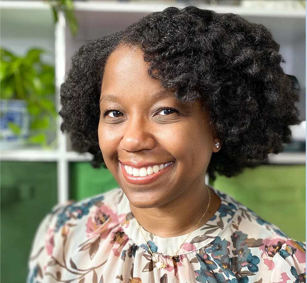 Picture of a smiling person with a long curly afro, dark skin, and a floral printed shirt against a green background.