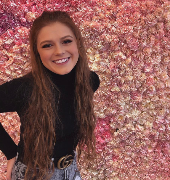 Picture of a smiling person with long strawberry blonde straight hair, fair skin, a black shirt and blue jeans against a pink background.