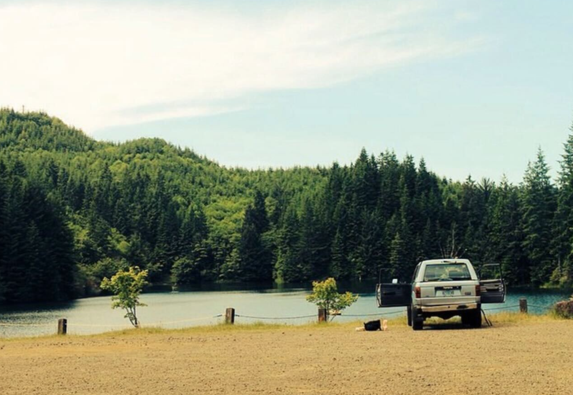 A picture of a lake with trees in the background, sand in the foreground and a Jeep with doors open on the right.