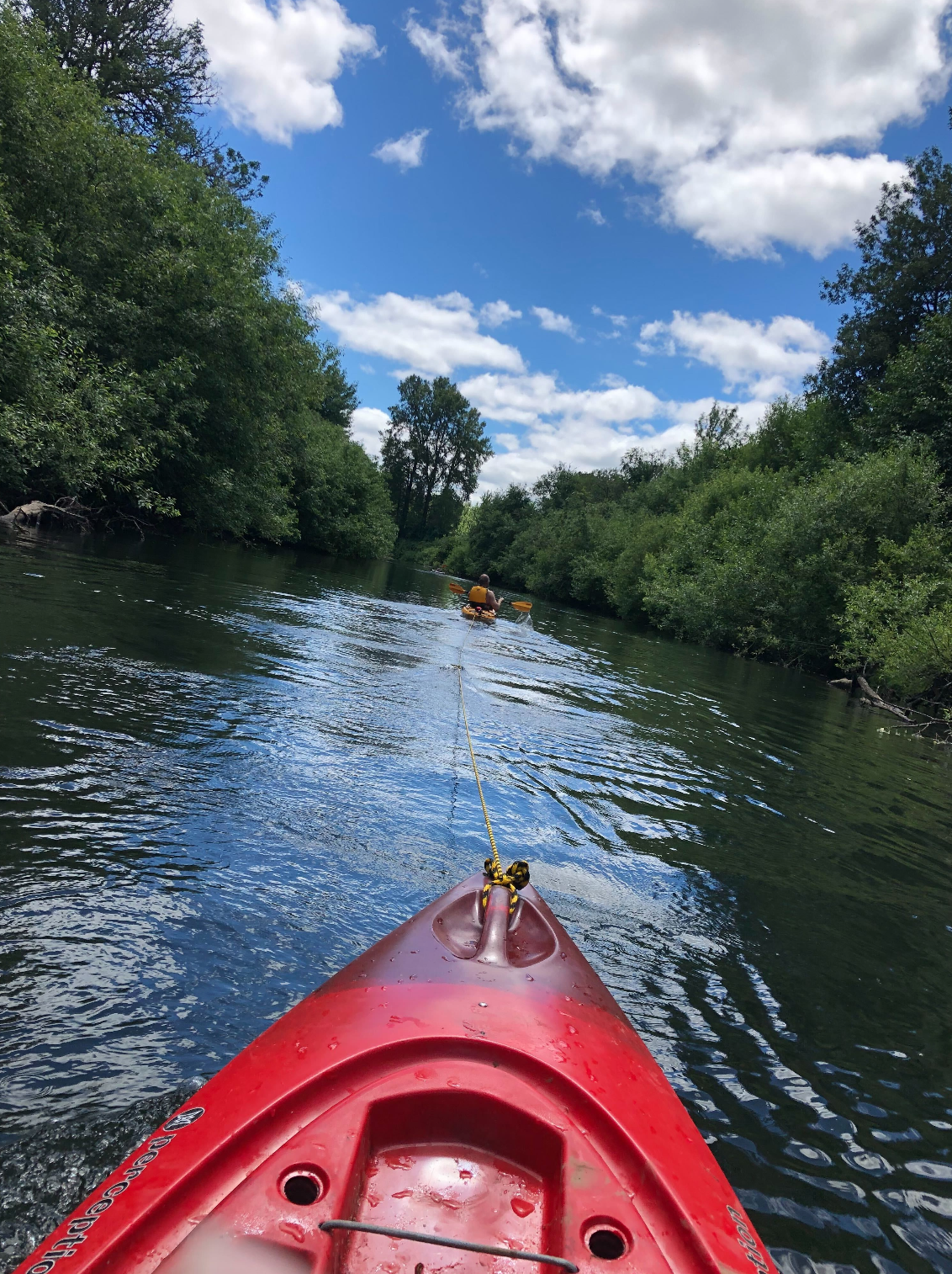 A picture from the inside of a red kayak on a river lined with trees with another person and kayak in the distance ahead. The kayaks are connected with a rope and the sky has some fluffy clouds.