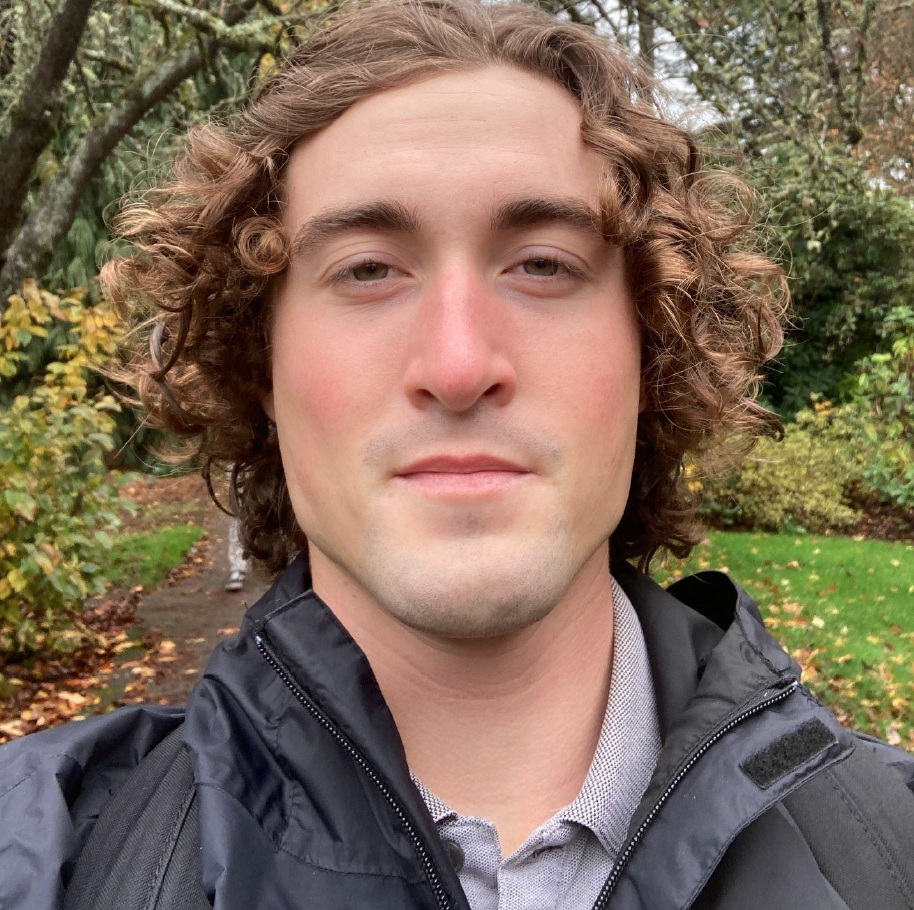 Selfie of a person with medium length curly hair, fair skin, and a black rain jacket against a background full of trees.