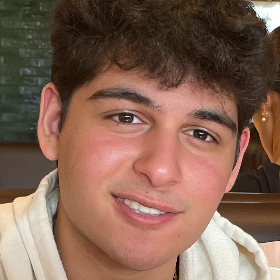 Selfie of a person with medium length curly hair, fair skin, and a black rain jacket against a background full of trees.