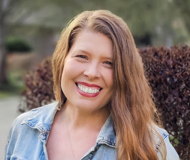 Picture of a smiling person with long strawberry blonde straight hair, fair skin, a black shirt and blue jeans against a pink background.